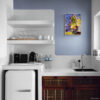 Botanical wall art in white frame displayed on blue accent wall in compact kitchen with white countertops and dark wood cabinets. Impressionist style potted plant painting with vibrant purple and yellow background. Modern kitchenette decor with retro white mini fridge, open shelving, and minimalist design. Perfect for small apartment kitchen, coffee station, or breakfast nook in contemporary or Mediterranean style home.