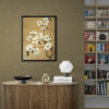 Elegant floral wall art in black frame hanging above mid-century modern wooden cabinet in sophisticated living space. White blossom painting on warm tan background complements olive-toned textured wall. Contemporary styling features ribbed wooden credenza with modern table lamp, decorative bowl, and stacked books. Built-in bookshelf visible adjacent to the vignette creating a refined, literary atmosphere.