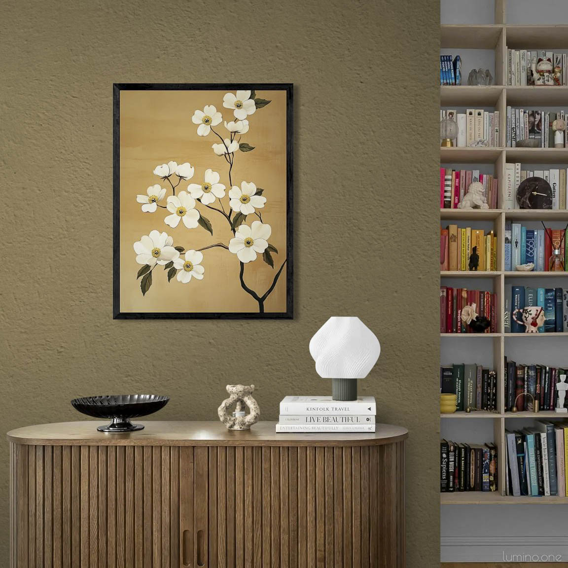 Elegant floral wall art in black frame hanging above mid-century modern wooden cabinet in sophisticated living space. White blossom painting on warm tan background complements olive-toned textured wall. Contemporary styling features ribbed wooden credenza with modern table lamp, decorative bowl, and stacked books. Built-in bookshelf visible adjacent to the vignette creating a refined, literary atmosphere.