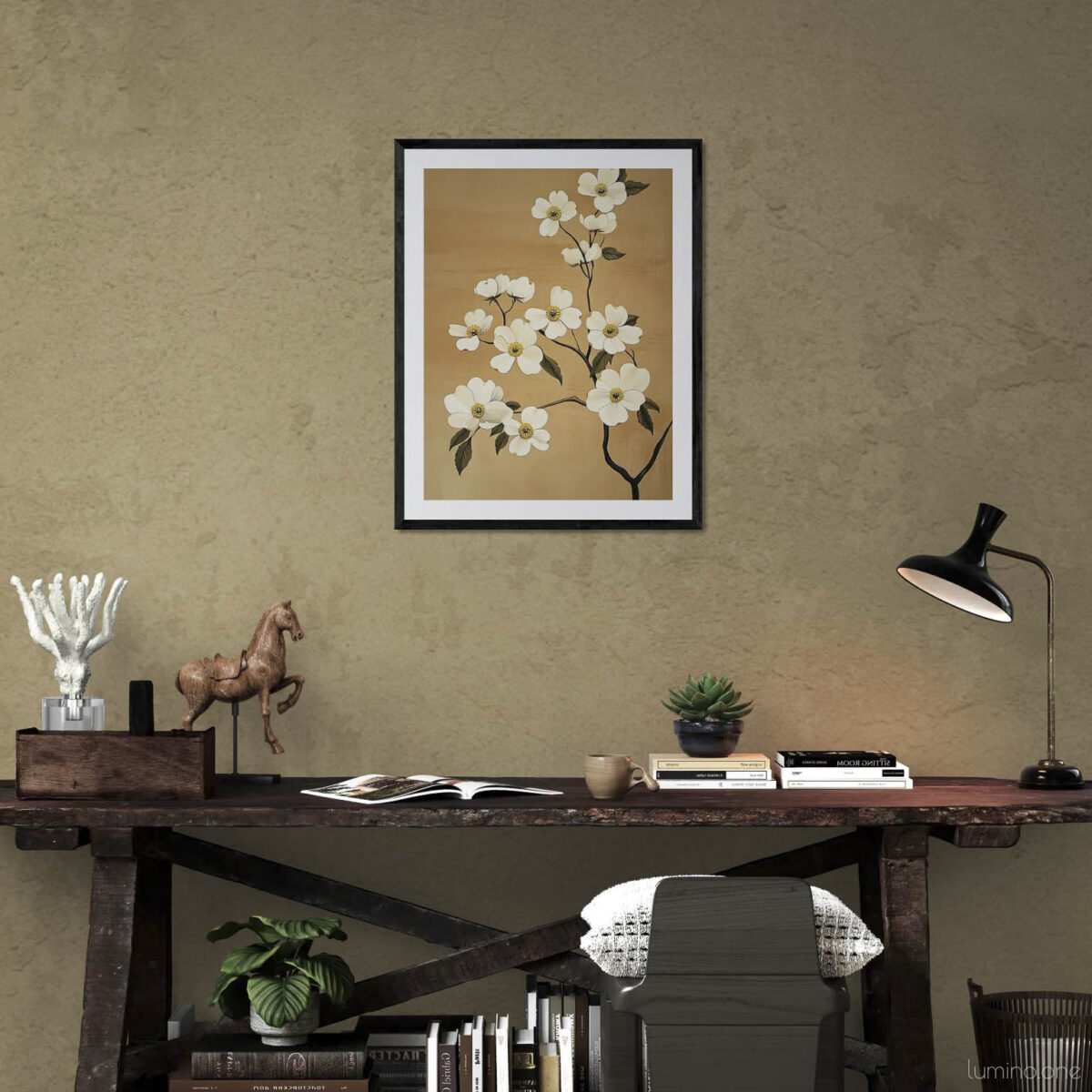 Elegant white floral art print in black frame hanging above rustic wooden desk in modern home office. Neutral taupe walls complement the warm tan background of the botanical artwork. Workspace features minimalist desk styling with vintage lamp, wooden horse figurine, small succulent plant, stacked books, and gray chair creating a sophisticated yet cozy work environment.