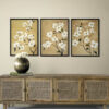 Set of three botanical wall art prints with white flowers on warm tan backgrounds displayed above rattan credenza in minimalist living room. Triptych floral artwork in black frames creates elegant focal point on cream wall. Natural wood media console with cane webbing doors styled with metallic round vase and white flowers for modern bohemian aesthetic.