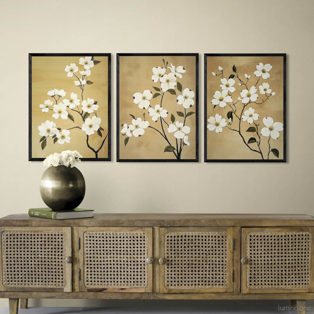Set of three botanical wall art prints with white flowers on warm tan backgrounds displayed above rattan credenza in minimalist living room. Triptych floral artwork in black frames creates elegant focal point on cream wall. Natural wood media console with cane webbing doors styled with metallic round vase and white flowers for modern bohemian aesthetic.