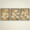 Set of three botanical wall art prints featuring white dogwood flowers on warm tan backgrounds in sleek black frames. Elegant floral triptych displayed against neutral cream wall showing detailed branch arrangements with delicate white blossoms and dark green leaves. Coordinated artwork set creates harmonious focal point with varying compositions of the same botanical theme.