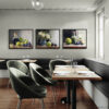 Three framed still life oil painting prints displayed as gallery wall in elegant restaurant dining room featuring fruits, flowers in blue vase, and glass bottles in classic painterly style, shown above green velvet banquette seating with wooden tables