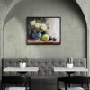 Framed still life oil painting print featuring white roses in a blue ceramic vase with fresh green apple and dark plums on wooden table, displayed in modern cafe or restaurant with arched alcove, gray banquette seating, and white bistro tables