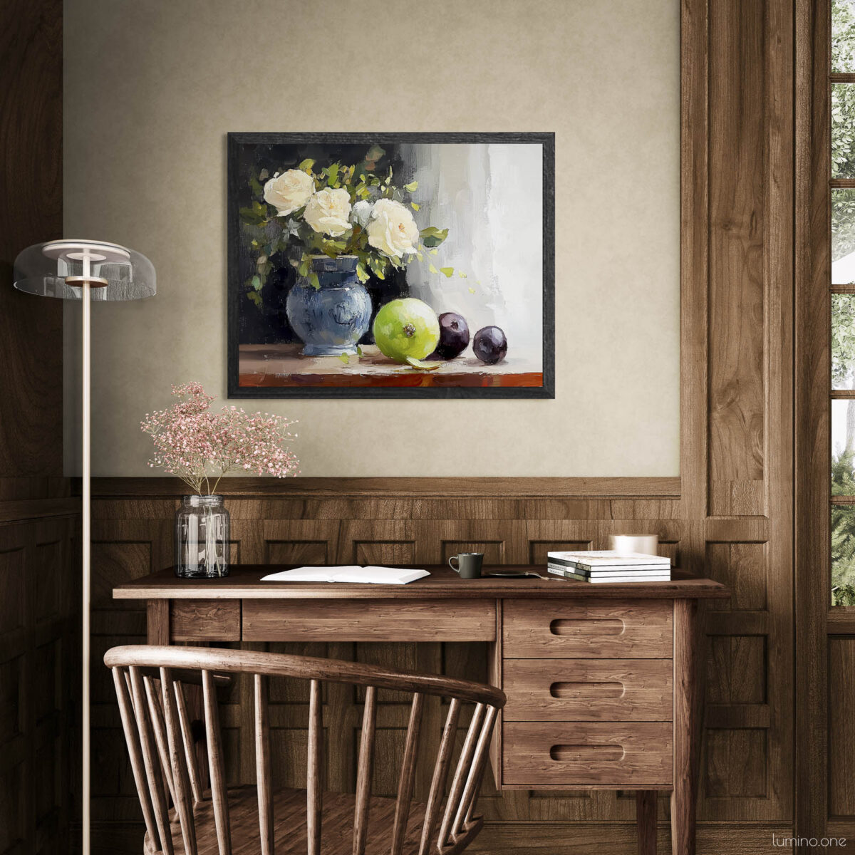 Framed still life oil painting print featuring white roses in a blue ceramic vase with fresh green apple and dark plums on wooden table, displayed above wooden desk in warm neutral home office setting