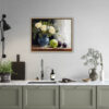 Framed still life oil painting print featuring white roses in a blue ceramic vase with fresh green apple and dark plums on wooden table, displayed above modern kitchen counter with sage green cabinets, marble countertop, and natural wood frame