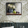 Framed still life oil painting print featuring white roses in a blue ceramic vase with fresh green apple and dark plums on wooden table, displayed in traditional dining room with sage green floral wallpaper, dark wood wainscoting, and brass chair with round table
