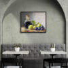 Framed still life oil painting print featuring green grapes, dark wine bottle with cork, and blue grapes arranged on wooden surface, displayed in modern cafe or restaurant with arched alcove, gray banquette seating, and white bistro tables