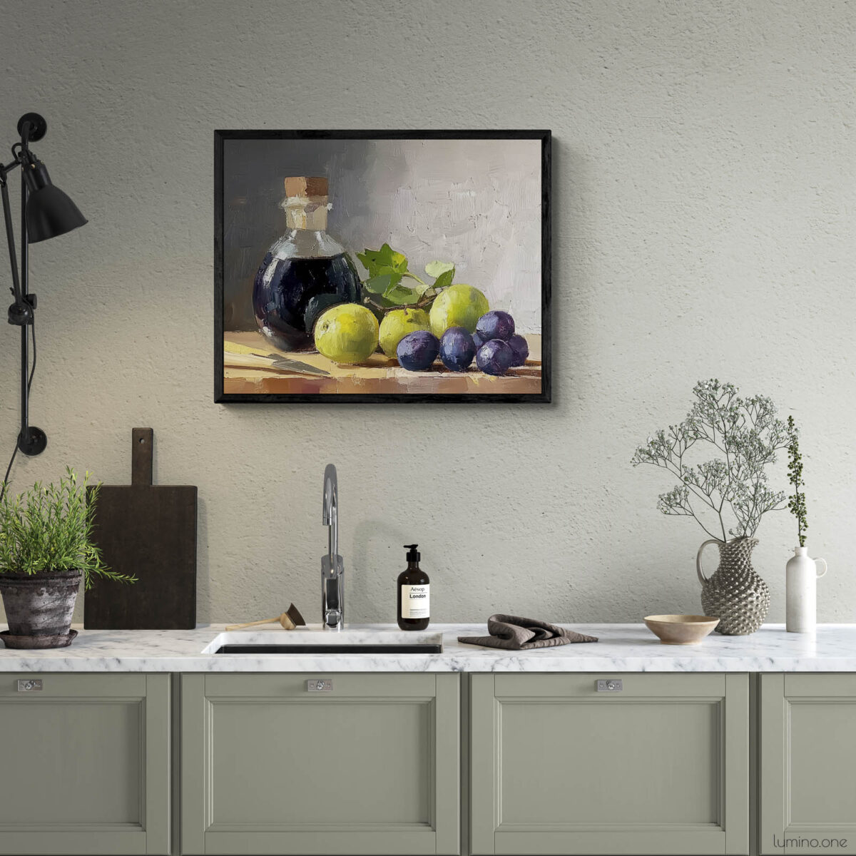 Framed still life oil painting print featuring green grapes, dark wine bottle with cork, and blue grapes arranged on wooden surface, displayed above modern kitchen counter with sage green cabinets and marble countertop