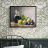 Framed still life oil painting print featuring green grapes, dark wine bottle with cork, and blue grapes arranged on wooden surface, displayed in traditional dining room with sage green floral wallpaper, dark wood wainscoting, and brass chair with round table