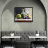 Framed still life oil painting print featuring green apple, dark plums, and green glass bottle with cork arranged on white cloth, displayed in modern cafe or restaurant with arched alcove, gray banquette seating, and white bistro tables