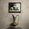 Framed still life oil painting print featuring green apple, dark plums, and green glass bottle with cork arranged on white cloth, displayed above antique black chest with ornate brass pitcher in rustic interior