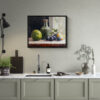 Framed still life oil painting print featuring green apple, dark plums, and green glass bottle with cork arranged on white cloth, displayed above modern kitchen counter with sage green cabinets and marble countertop