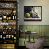 Framed still life oil painting print featuring green apple, dark plums, and green glass bottle with cork arranged on white cloth, displayed in wine bar or restaurant with exposed brick wine storage shelves, green banquette seating, and purple velvet chairs