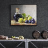 Framed still life oil painting print featuring green grapes, dark wine bottle with cork, and blue grapes arranged on wooden surface, displayed in wine cellar or bar area with dark textured walls above industrial metal shelving with cheese board and wine barrels
