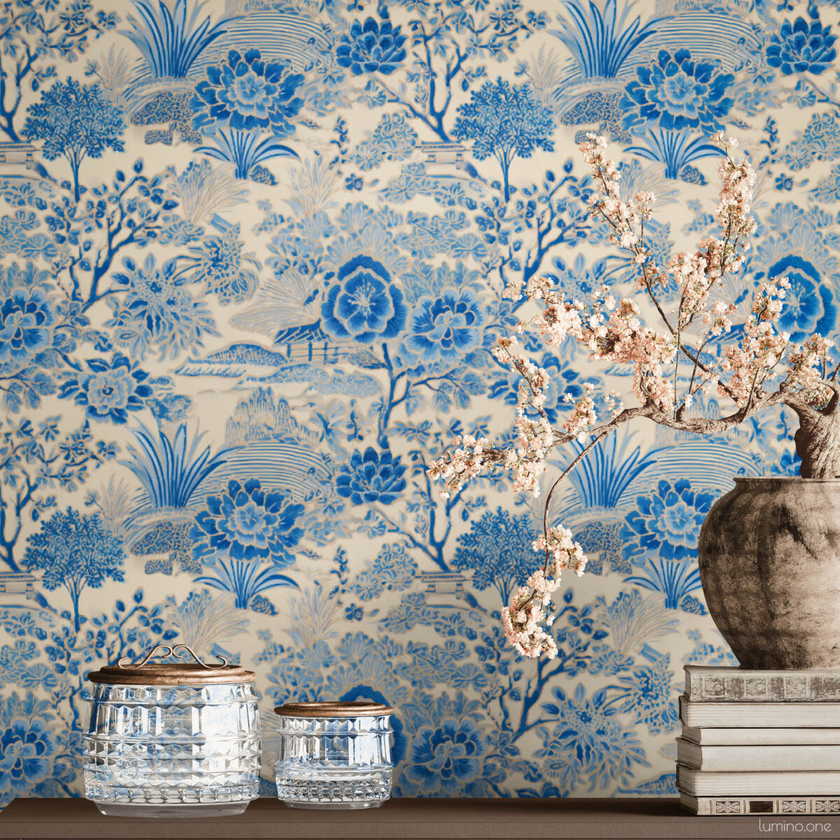 Blue and white toile peel and stick wallpaper on a full wall behind a taupe modern sideboard, featuring a vintage botanical garden pattern with large peony flowers and trees. Styled with a rustic stoneware vase holding cherry blossom branches, stacked vintage books, and crystal glass jars with brass lids.