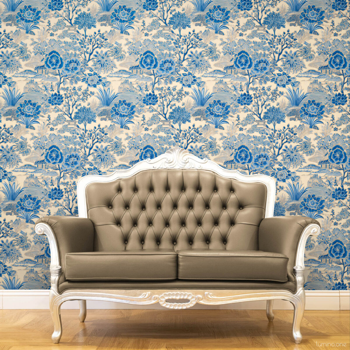 Blue and white toile peel and stick wallpaper covering a full wall behind an ornate French-style tufted leather loveseat with silver carved wood frame, featuring a vintage botanical garden pattern with large peony flowers and trees. Styled on a warm honey herringbone wood floor, showcasing a classic French country living room aesthetic.