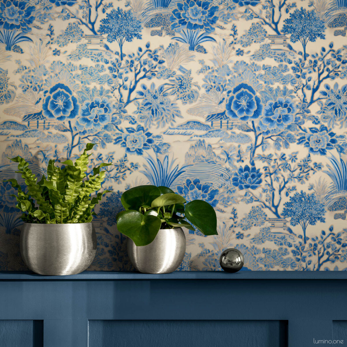 Blue and white toile peel and stick wallpaper above navy blue painted wall paneling, featuring a vintage botanical garden pattern with flowers and trees. Styled with two round brushed silver planters holding a crispy wave fern and a pilea plant, with a small chrome decorative sphere, showing how the wallpaper pairs beautifully with blue wainscoting and modern metallic accents.