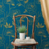 Dark teal and gold peony removable wallpaper styled as a cozy reading vignette with a vintage bentwood chair holding stacked art books, a face planter with grass, and a flowing gold curtain