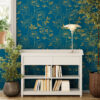 Bright entryway accent wall with dark teal and gold peony peel and stick wallpaper, styled with a white console bookshelf, silver table lamp, and lush indoor plants on wood floors